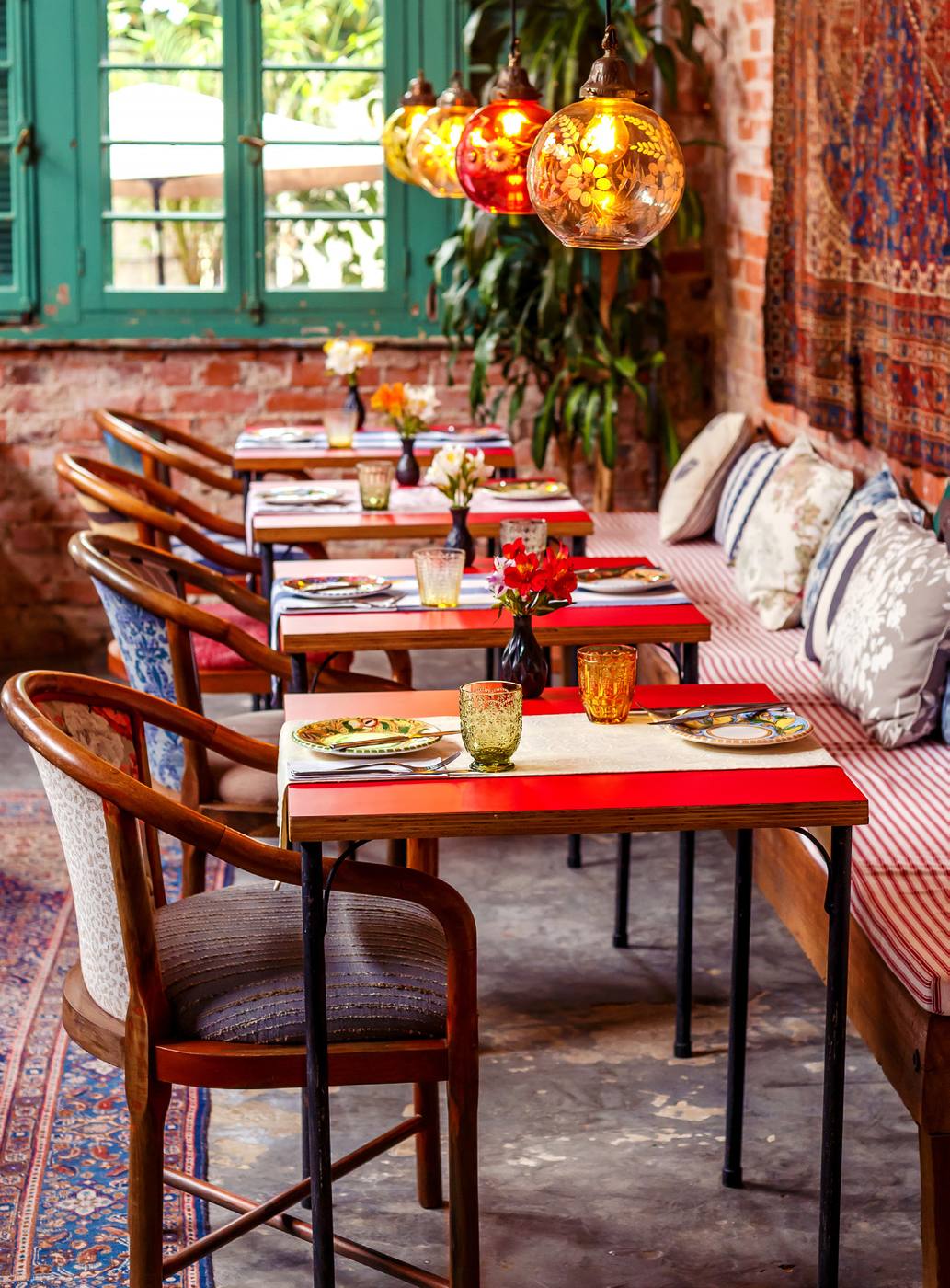Salão interno do Aora Cucina com tijolo à vista, tapetes persas, luminárias de vidro colorido e mesas postas