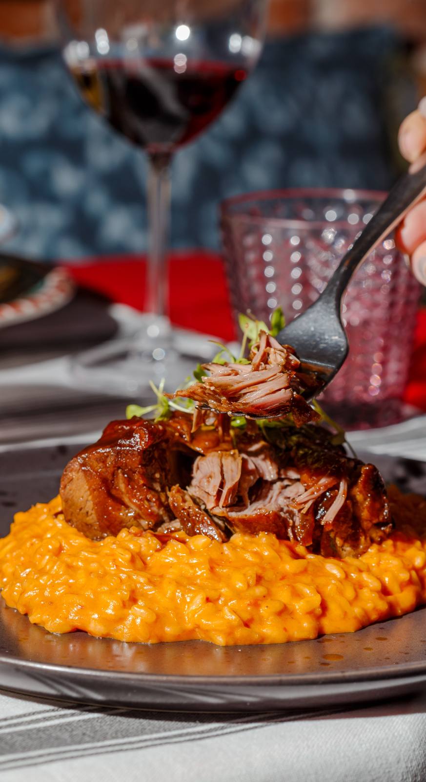 Paleta de cordeiro braseada servida sobre risoto de tomate, com garfo erguendo um pedaço da carne